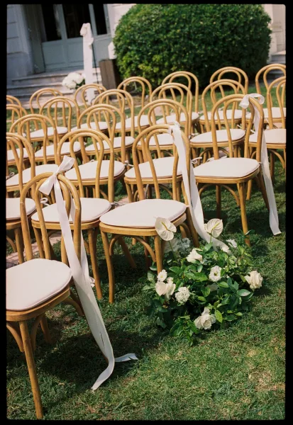Ceremony seating with bentwood wedding chairs and white ribbons line a grass lawn aisle before a building facade with steps and doors.