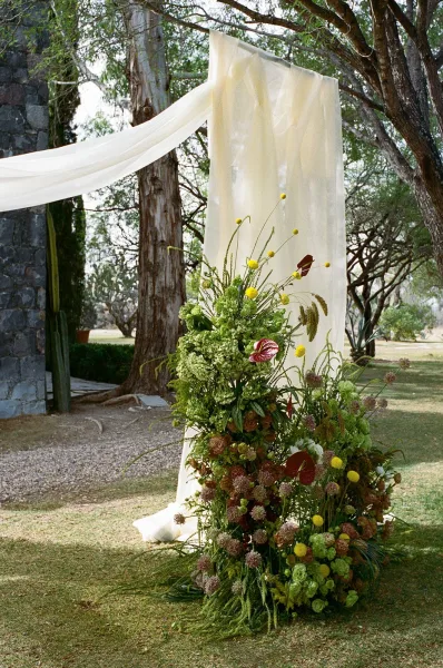 Wedding ceremony altar with sheer drapery on a wooden arch frame and grounded floral arrangement on a lawn beside a stone wall