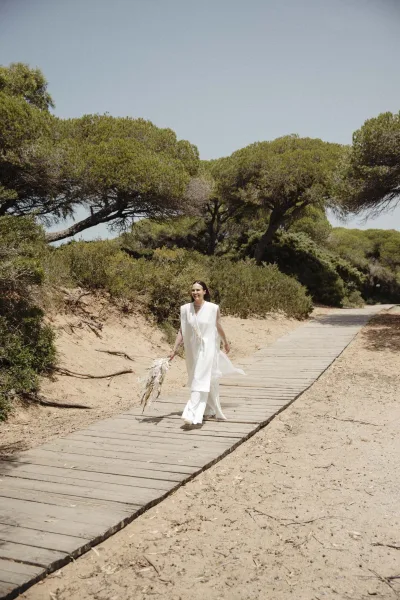 Bridal portrait of a bride walking with bouquet in a minimalist white gown and sandals on a boardwalk by sand dunes under blue sky