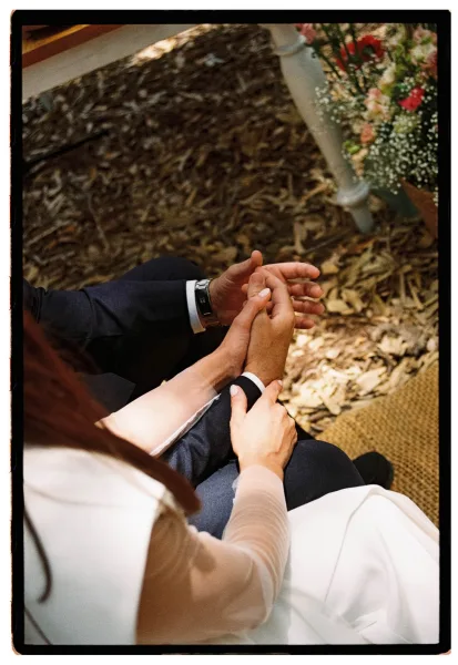 Ceremony handholding close-up of bride and groom, long-sleeve gown and groom’s wristwatch beside bouquet ribbon over wood-chip aisle