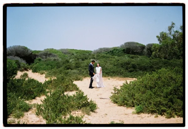 Couple portrait with bride in minimalist gown and long veil holding a pampas bouquet beside groom in black tux on sand dunes
