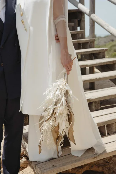 Wedding couple portrait of bride and groom standing on rustic wooden stairs, bride in cape holding a dried foliage bouquet outdoors