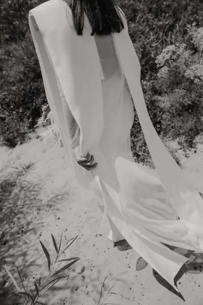 Bridal gown details on a long sleeve wedding dress with a flowing train, seen from behind as the bride walks along a sandy path by shrubs