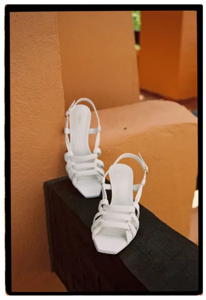 Bridal shoes, white wedding heels with ankle straps and buckles resting on a wooden ledge against a warm terracotta stucco wall