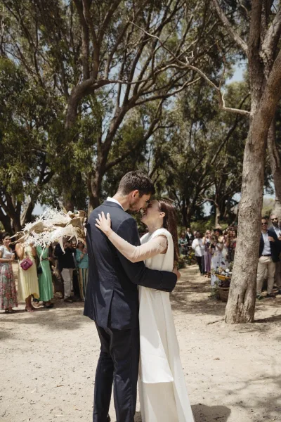 Ceremony kiss as bride in long sleeve gown and groom in navy suit embrace, holding a dried bouquet on a tree-lined path with guests watching