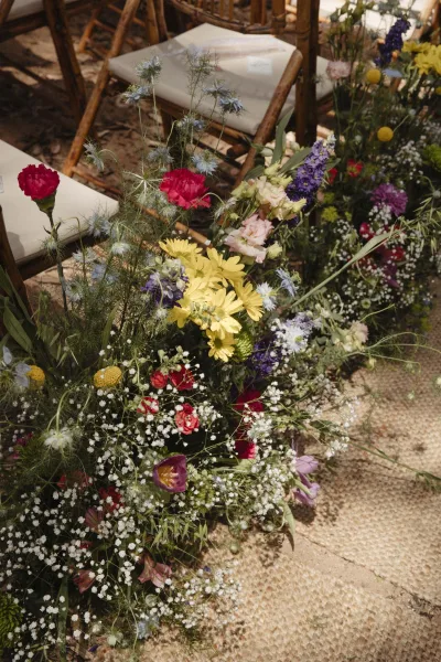 Ceremony aisle flowers in a wildflower aisle arrangement with baby’s breath, yellow daisies, and purple blooms along a dirt path beside wooden chairs