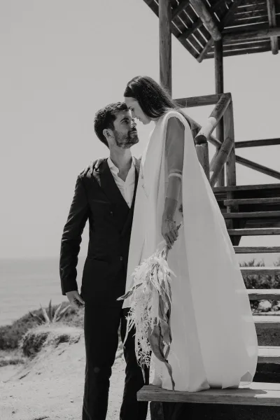 Couple portrait in a black and white wedding portrait, bride in bridal cape holding bouquet as they touch foreheads by beach stairs