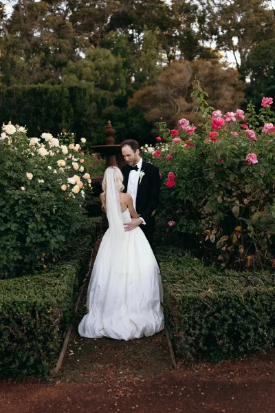 Couple portrait as the bride and groom embrace along a garden path, her veil catching light by the fountain amid rose bushes and hedges.