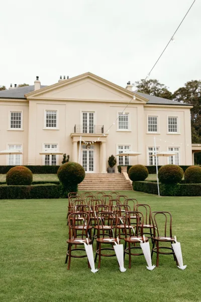 Ceremony seating arranged for an outdoor ceremony setup with bentwood chairs on a lawn before a mansion facade and topiary hedges.