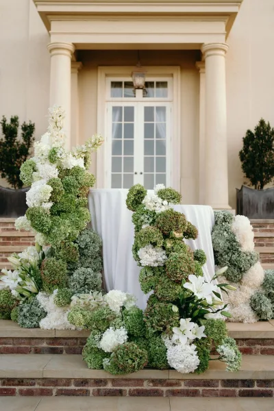 Ceremony setup featuring wedding altar flowers in white hydrangeas, arranged on stone steps before columns and French doors of the building facade.