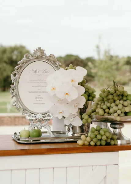 Bar menu sign, a signature cocktail sign in a silver frame, displayed outdoors against garden trees and a warm brick wall.