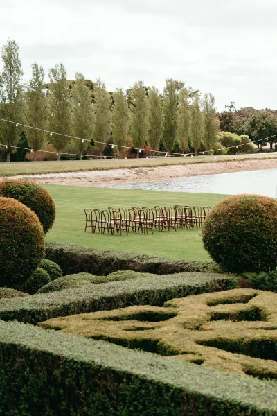 Ceremony seating with outdoor ceremony chairs in rows on a grass lawn, under string lights beside a pond and topiary hedges.