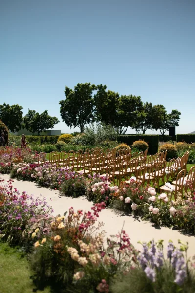 Ceremony aisle design for an outdoor ceremony aisle lined with pastel florals on a grass lawn framed by garden hedges and trees under a blue sky.