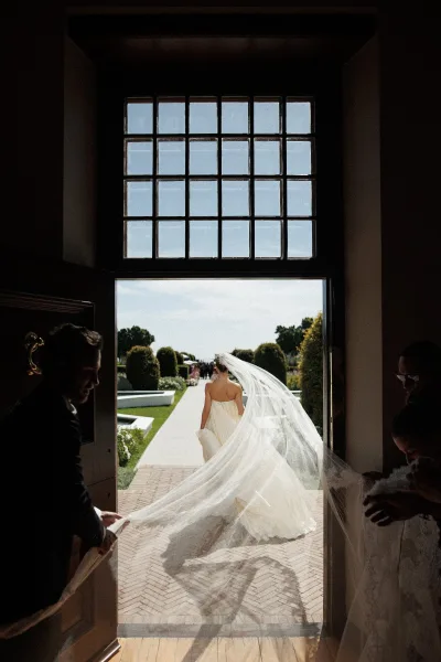 Bride entrance, walking down the aisle with a flowing bridal veil along a brick walkway between garden hedges under open sky.