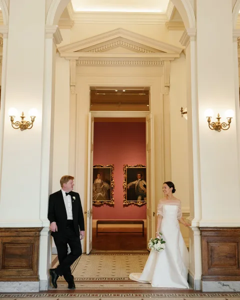 Couple portrait of bride holding bouquet beside groom in black tuxedo, standing in a grand hallway with arched doorway and tile floor