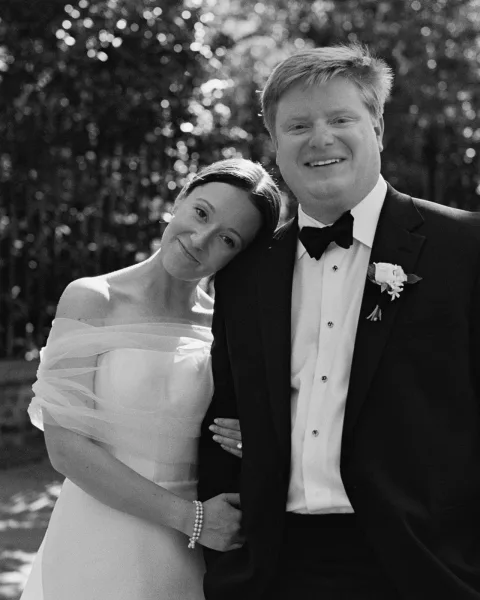 Couple portrait in a black and white wedding portrait style, bride leaning on groom in tuxedo amid sunlit trees and greenery bokeh