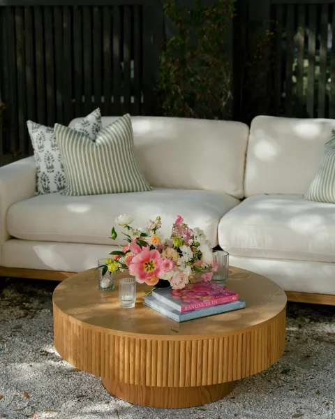Lounge seating vignette in a wedding lounge area with white outdoor sofa, striped pillows, floral centerpiece and votives on a low wooden table in dappled sun