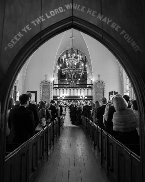 Ceremony moment at a church wedding ceremony, bride in gown with long veil walking the wooden aisle toward the altar as guests watch
