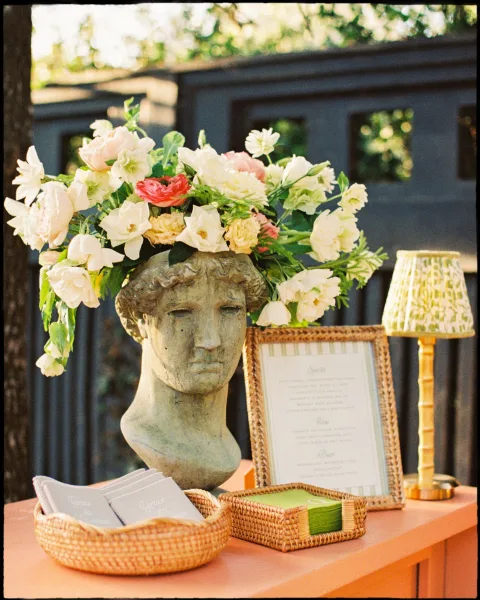 Wedding welcome table with a wedding welcome sign display, floral arrangement in bust vase, wicker baskets, and lamp set against garden greenery