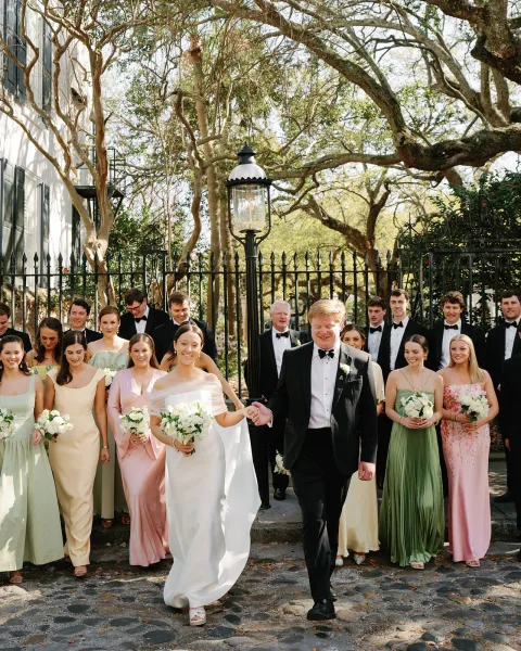 Wedding party portrait with bride and groom walking, holding bouquets beside a wrought iron fence on a stone path outside a white building