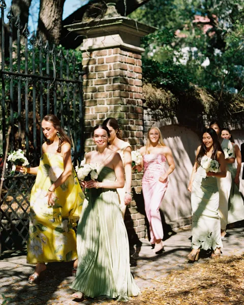 Bridesmaid processional with bridesmaids walking down aisle in mismatched dresses, holding white bouquets by a wrought iron gate in sunlight
