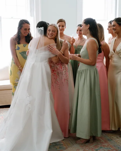 Bride with bridesmaids hugging bride in her veil and wedding dress, joyful reveal in bright window light against white walls