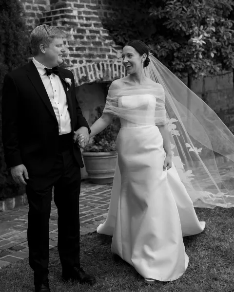 Couple portrait of bride and groom holding hands, bride laughing in veil and satin dress, walking by a brick wall with greenery