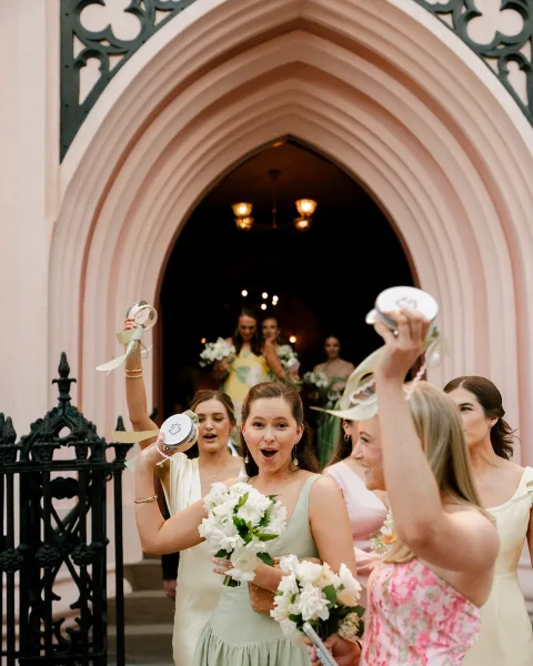 Wedding exit with bridesmaids cheering and waving ribbon wands, holding white bouquets and tambourines at a church doorway arch and gate