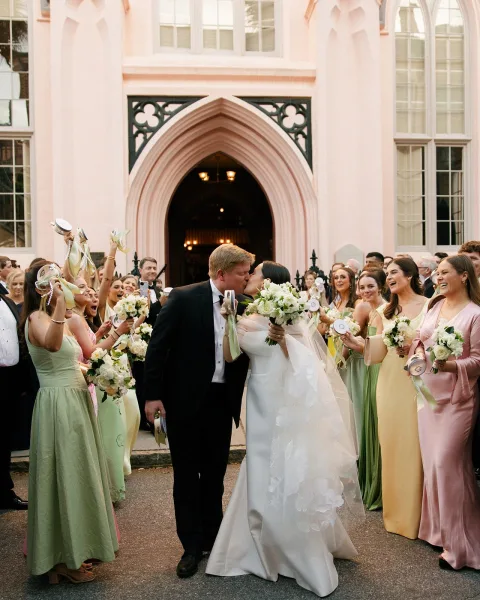 Wedding recessional as bride and groom kiss at a church entrance, bride in strapless dress and veil holding bouquet, guests cheering outside