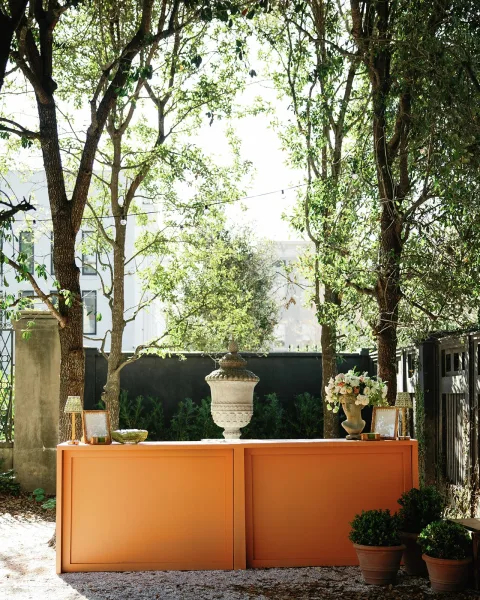 Wedding bar setup featuring an orange wedding bar with framed menu signs, table lamps, florals, and string lights in a garden setting