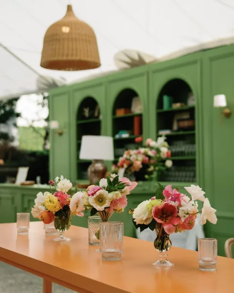 Reception tablescape with bud vase centerpiece of colorful wedding flowers, ribbed glass tumblers, and a lamp beneath green bookshelf backdrop.