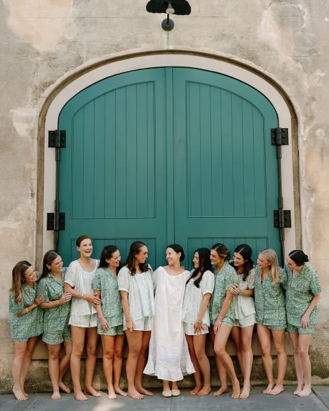Bridesmaid group photo with bridesmaids in matching pajamas gathered barefoot by an arched green door on a stone walkway, laughing