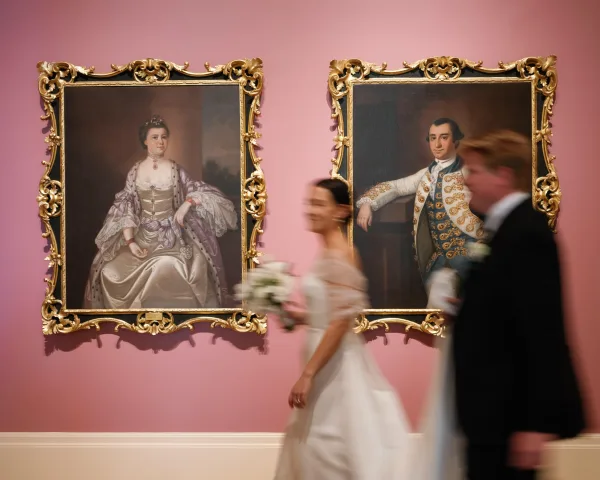 Wedding couple photo of bride and groom walking, bride holding bouquet, in an art gallery with framed paintings on a pink wall