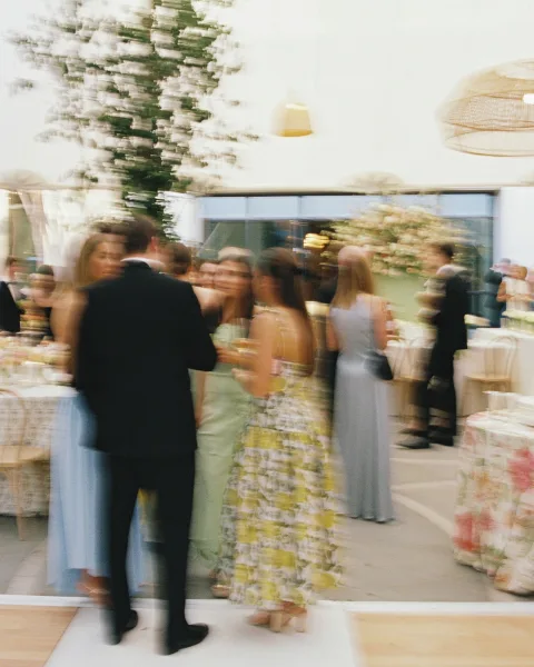 Wedding reception with guests mingling, holding champagne flutes around round tables with floral centerpieces under hanging pendant lights