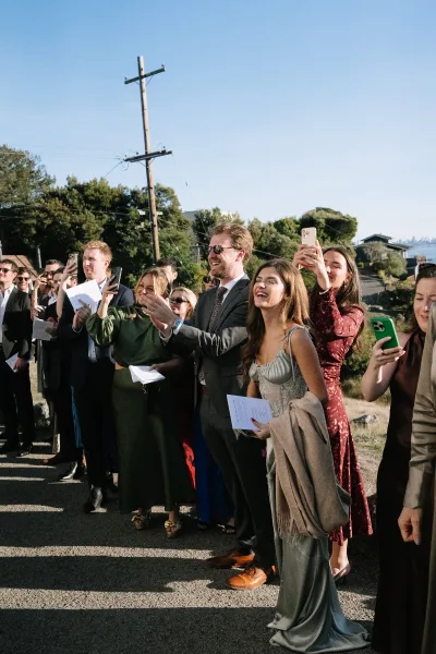 Wedding guests with lively guest reactions, some raising cell phones during an outdoor ceremony under a blue sky and trees.