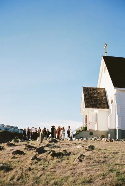 Church wedding exterior with a simple cross above the facade, set against blue sky and a rocky hillside with distant buildings.