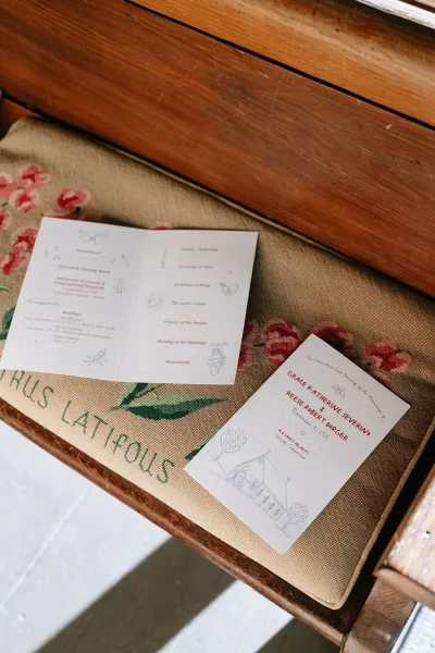 Wedding program, an order of service card featuring floral embroidery, resting on a wood pew against a gray floor.