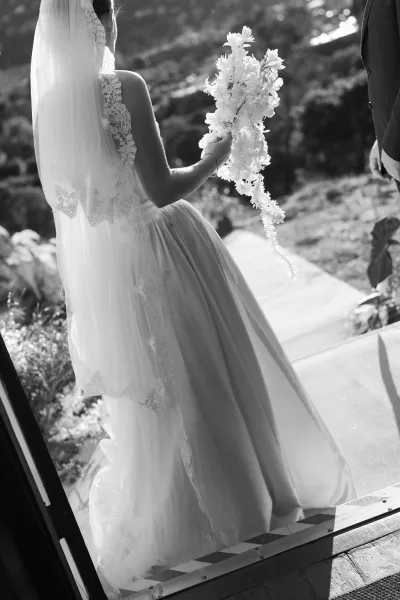 Bridal portrait of a bride holding bouquet beneath a lace veil on a garden walkway, framed by lush foliage and a softly rising hillside.