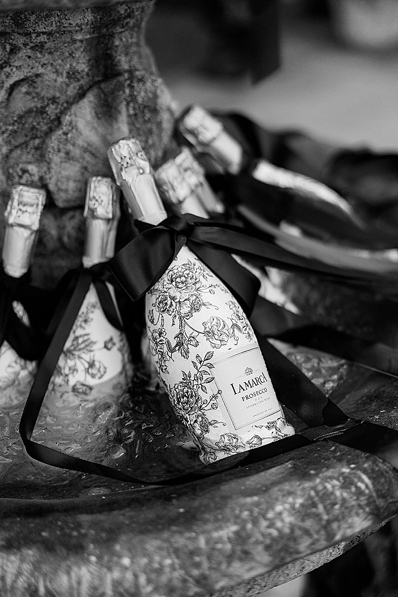 Champagne bottle detail with wedding champagne bottles tied in black ribbon bows and a floral label on a stone ledge against a wall
