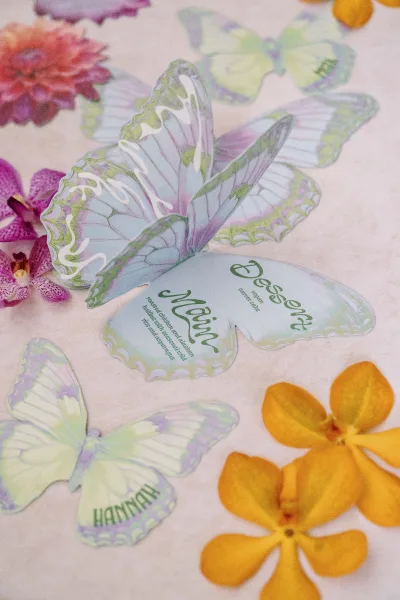 Wedding place cards with butterfly paper cutouts and printed names, styled with orchids and scattered petals on light table linen