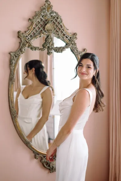 Bridal portrait of a bride by window light, looking over her shoulder in an ornate gold mirror against a pink wall with curtains