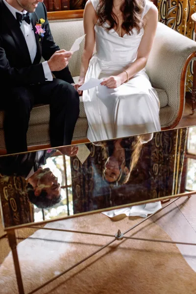 Wedding vows as couple reading vows on a vintage sofa, holding vow cards in window light beside a mirrored coffee table reflection