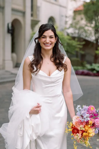 Bridal portrait of a smiling bride holding bouquet in a satin dress and long veil, standing on a courtyard walkway by white archways and greenery