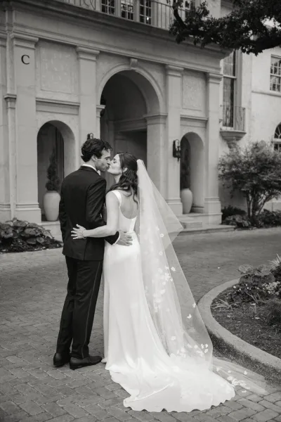 Wedding kiss portrait of bride and groom kiss, her long lace veil trailing as they embrace by an arched doorway and stone columns