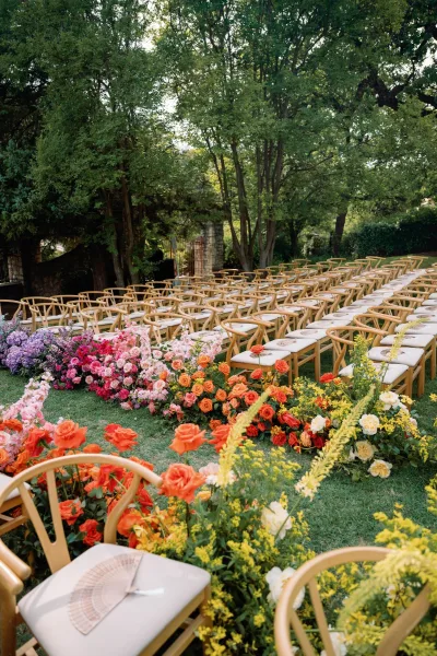 Outdoor ceremony setup with wood chairs and colorful rose aisle flowers, printed programs on seats, on a green lawn by trees and stone wall