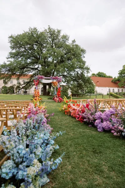Outdoor ceremony setup with a colorful floral arch and aisle flowers on a grass lawn, wooden chairs under a large tree by a white building