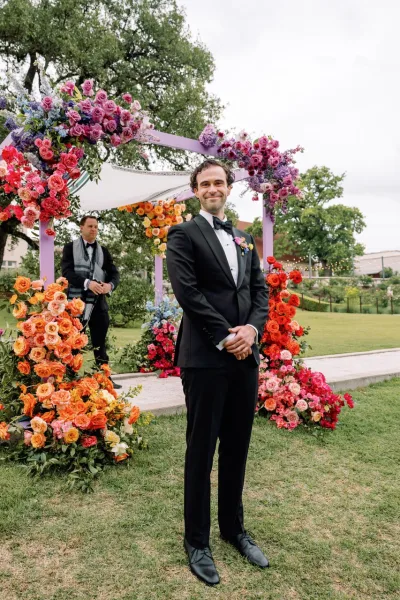 Groom portrait in a black tuxedo with bow tie and boutonniere, hands clasped before a colorful floral ceremony arch on a lawn