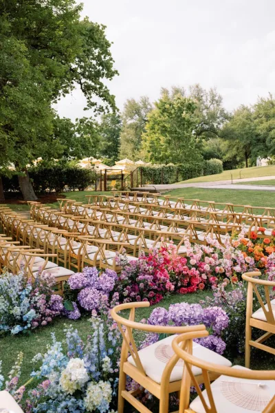 Ceremony aisle decor with flower lined wedding aisle of hydrangeas, roses, and greenery between wood chairs on a grassy lawn under clouds