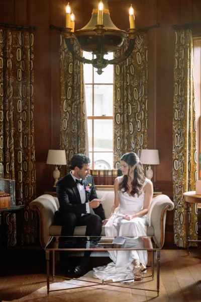 Couple portrait of bride and groom sitting on a vintage sofa as the groom reads a note, under a chandelier in a wood-paneled room