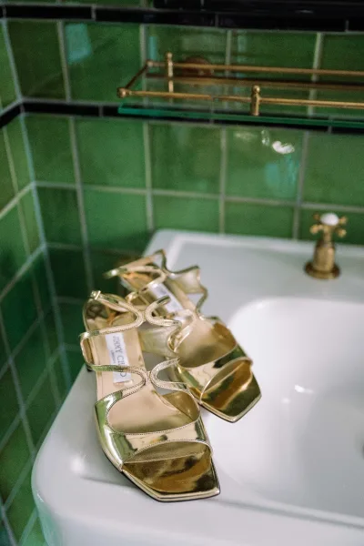 Wedding shoes, gold wedding heels resting on a bathroom sink beside a brass faucet against a distinctive green tile wall